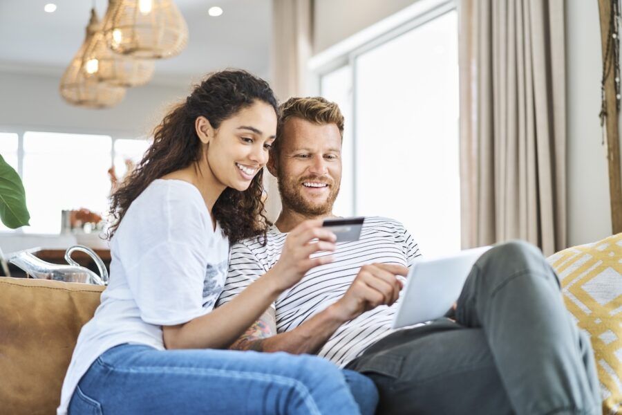 Woman sitting with credit card by man paying through electronic banking. Smiling multi-ethnic couple is enjoying shopping through digital tablet. They are in living room at home.