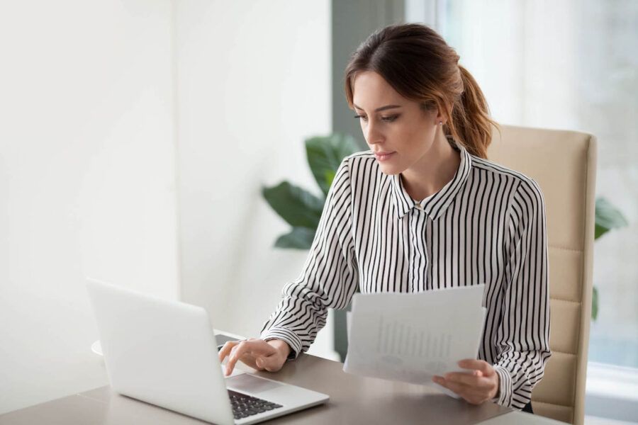 Serious focused businesswoman typing on laptop holding papers preparing report.