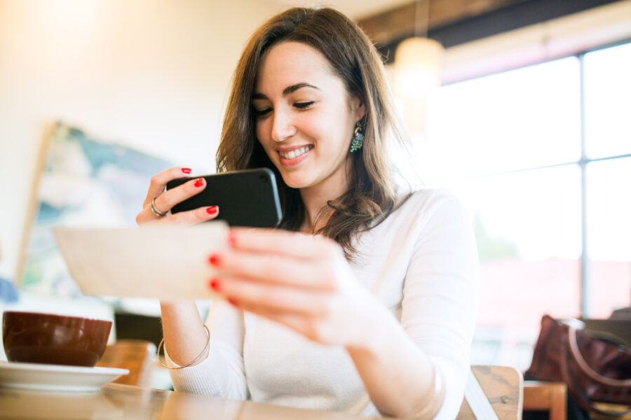 A smiling young woman takes a picture with her smart phone of a check or paycheck for digital electronic depositing, also known as