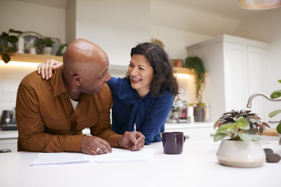 A Couple Reviewing And Signing Life Insurance Paperwork In Kitchen At Home.