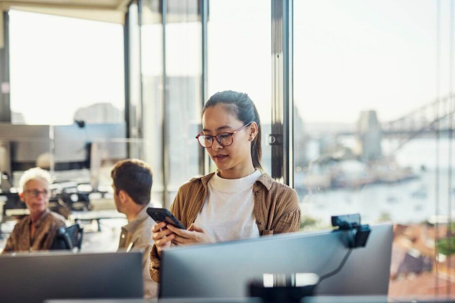 Woman canceling her travel plans over the phone due to a busy work week, standing in the modern office with colleagues in the background