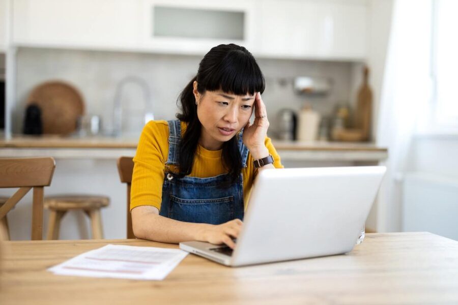 Concerned woman using her laptop in the kitchen, a few bills lying next to her on the table