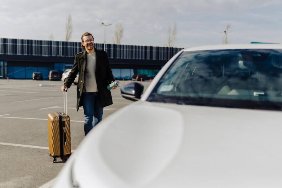 Smiling man with a suitcase and a water bottle approaching the rental car next to the airport