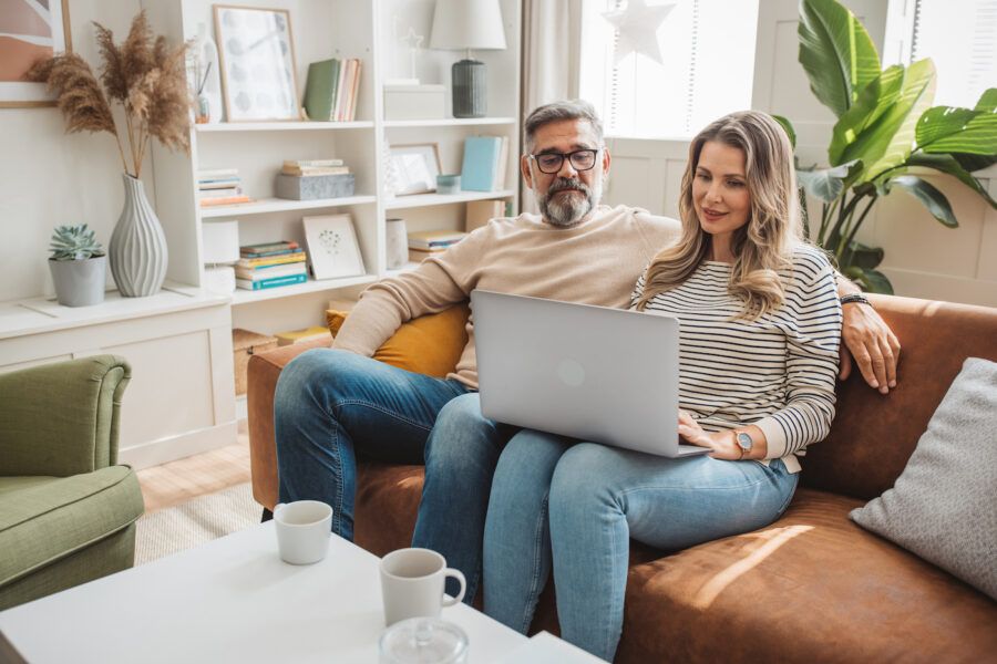 Middle aged couple sitting on sofa and using laptop.