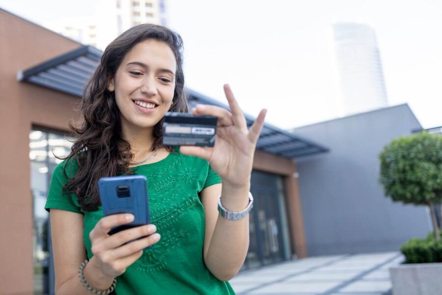 Smiling young woman looking at her credit card and holding a smartphone while standing outside of an office building
