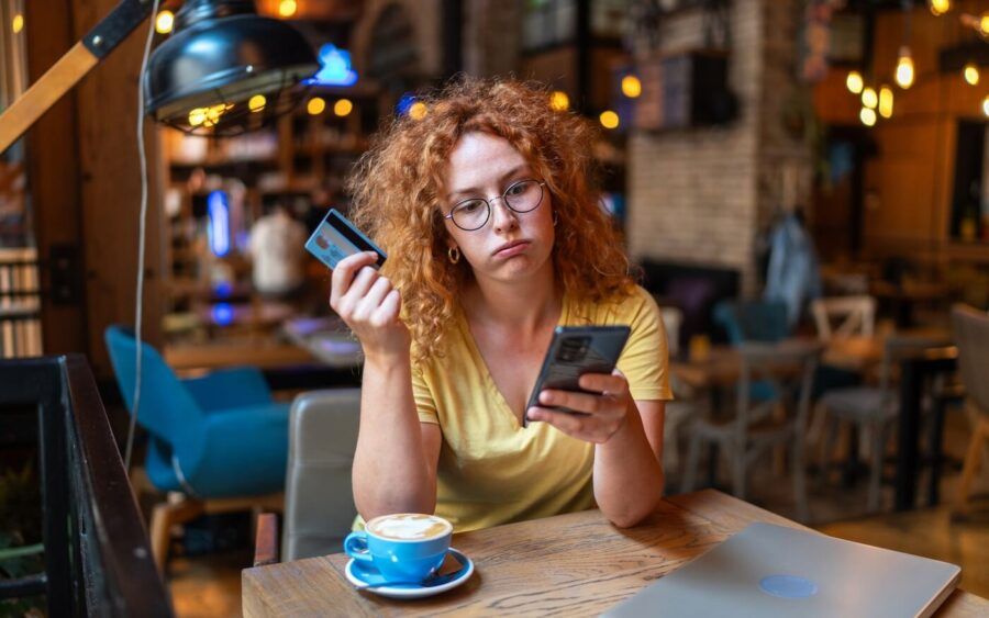 Upset young woman checking her smartphone while holding a credit card, sitting in a cozy cafe with a cup of coffee and a laptop