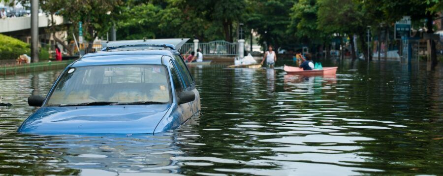 A blue car partially submerged in flood water on a tree-lined street.