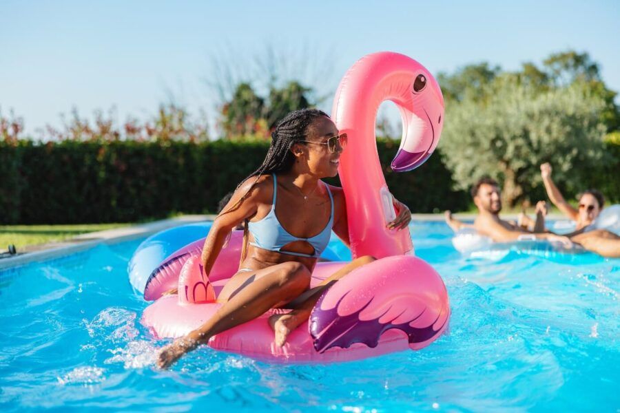 A happy woman enjoys a hot summer day in the pool with a giant inflatable flamingo, with her friends swimming in the background