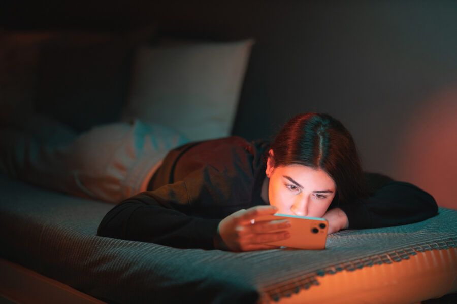 A teenage girl lies on the bed in her room lightened with orange and teal neon lights and updates her social media privacy settings on her mobile phone.