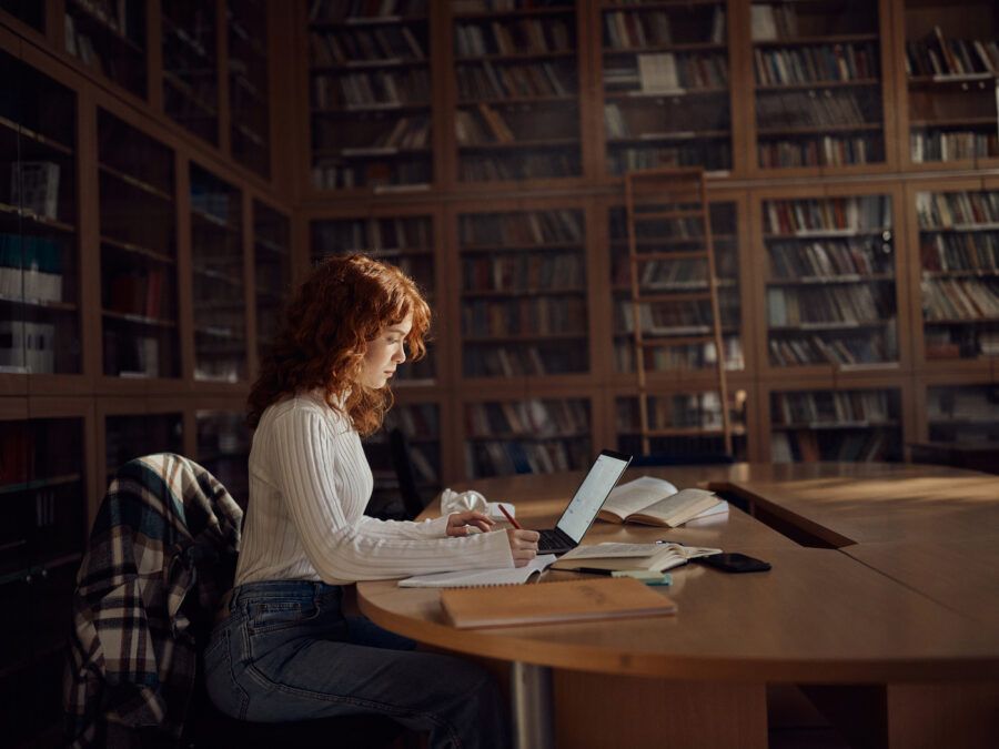A female student taking notes while using a computer in library.