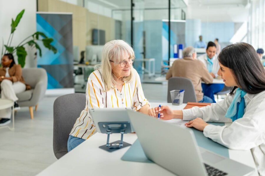 A female bank clerk assisting an elderly woman by explaining how to endorse a check, both seated at a desk in a bank branch.