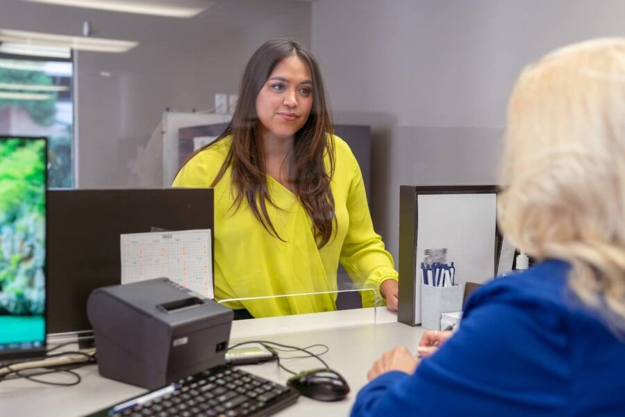 A young woman speaking with a female bank clerk through a glass counter window at a bank.