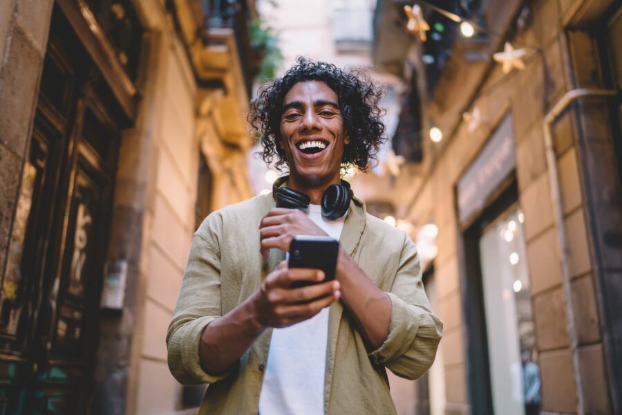 A smiling man in casual clothes with wireless headphones browsing smartphone while standing on narrow street between stone buildings in daytime.