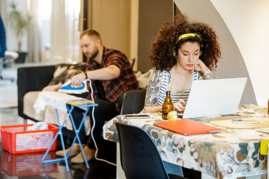 Young tattooed woman with headphones working on laptop at home, her flatmate ironing in the back