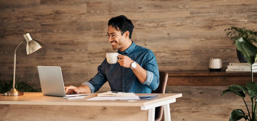 A happy man sitting at a desk and using his laptop to open a high yield savings account while holding a cup of coffee.