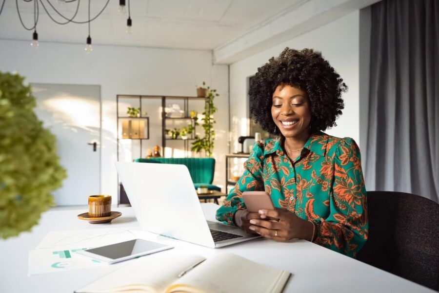 Smiling woman checking her credit score via smartphone while sitting at the desk with an open laptop, a tablet, and a notepad beside her