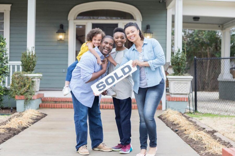 A happy family of four standing next to their new house and holding a 'sold' sign