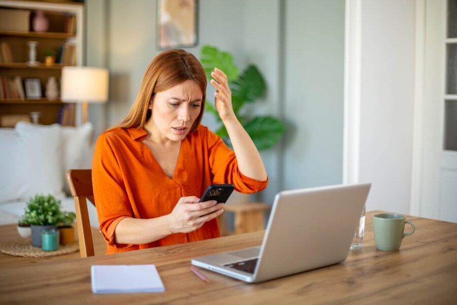 Frustrated woman looking at her mobile phone while sitting at the table with an open laptop and a notepad