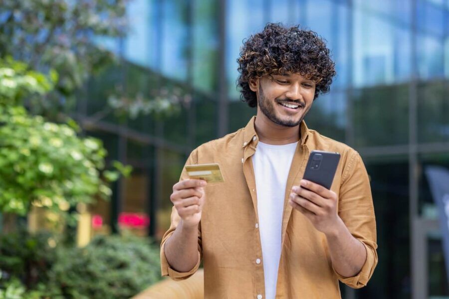 Smiling young man standing outdoors holding a credit card and a smartphone in front of a modern glass building