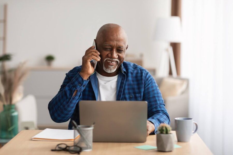 Smiling senior man making a phone call while looking at the laptop screen
