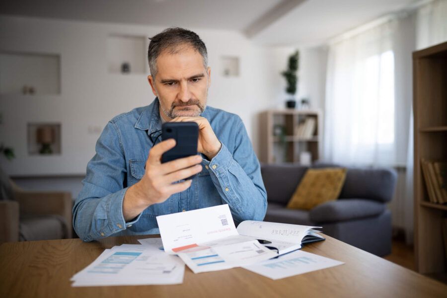 A middle-aged man sits at a wooden table, focused on his smartphone. Surrounding him are various documents and bills, indicating he is reviewing important information. The setting is a cozy living room with natural light coming through the window.