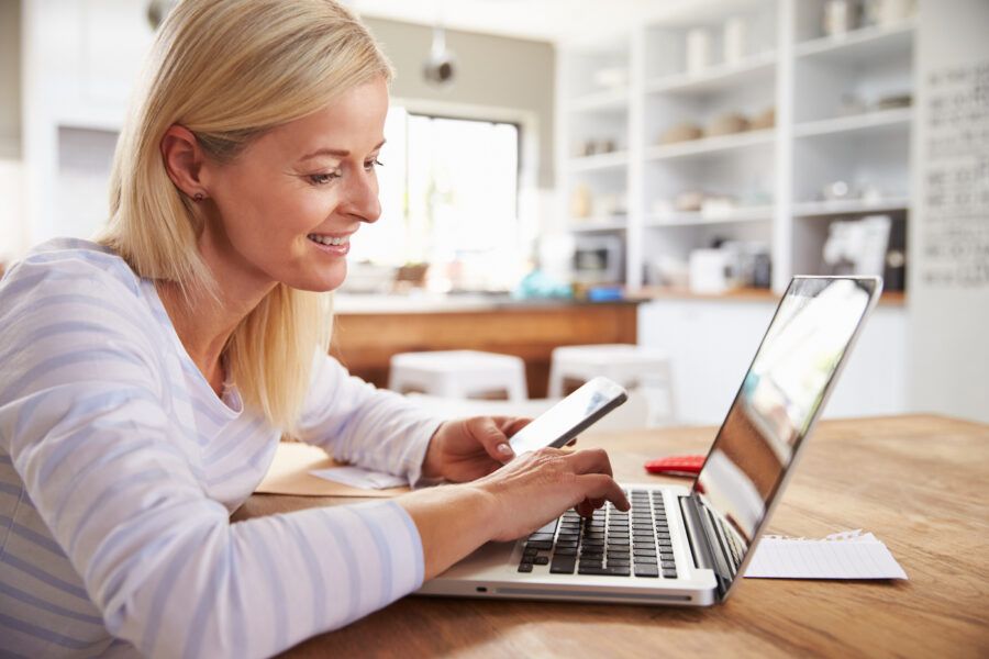 Woman working on laptop at home, holding a mobile phone