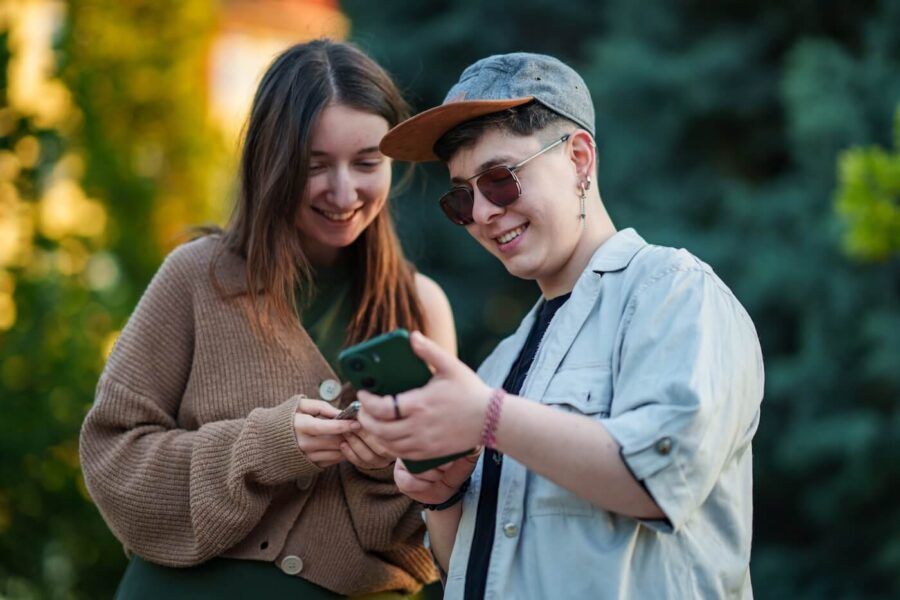 Two people standing outdoors and looking at a smartphone together. Trees and greenery create a blurred background, while both individuals focus on the mobile device.