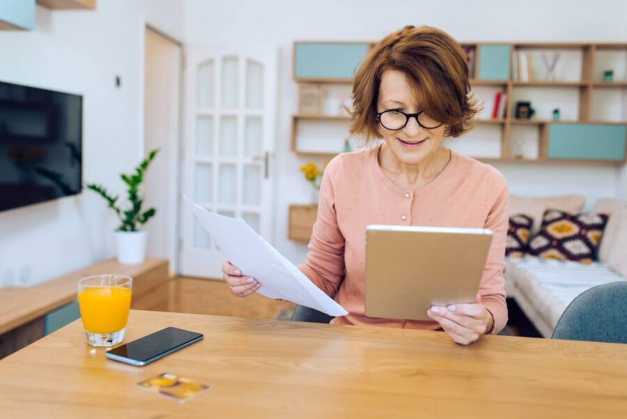 Focused mature woman comparing printouts with the data on her tablet at home
