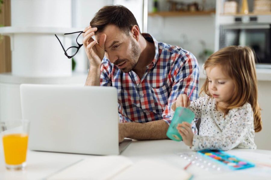 Concerned man took off his glasses and is looking at the laptop screen, while a small girl is painting with watercolor next to him