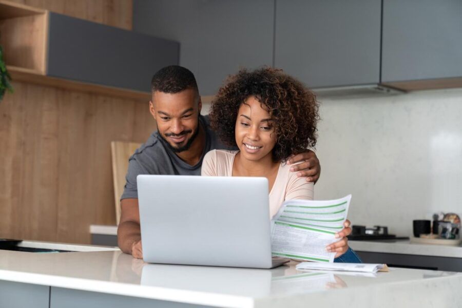 Smiling couple sitting together at home, using a laptop to plan their budget; the woman is holding printed documents.