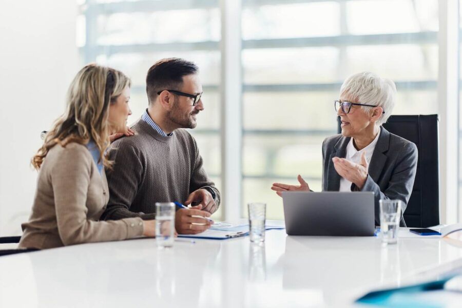 A couple sitting together with a female financial advisor in a bright office setting, reviewing documents, with a laptop and glasses of water on the table.
