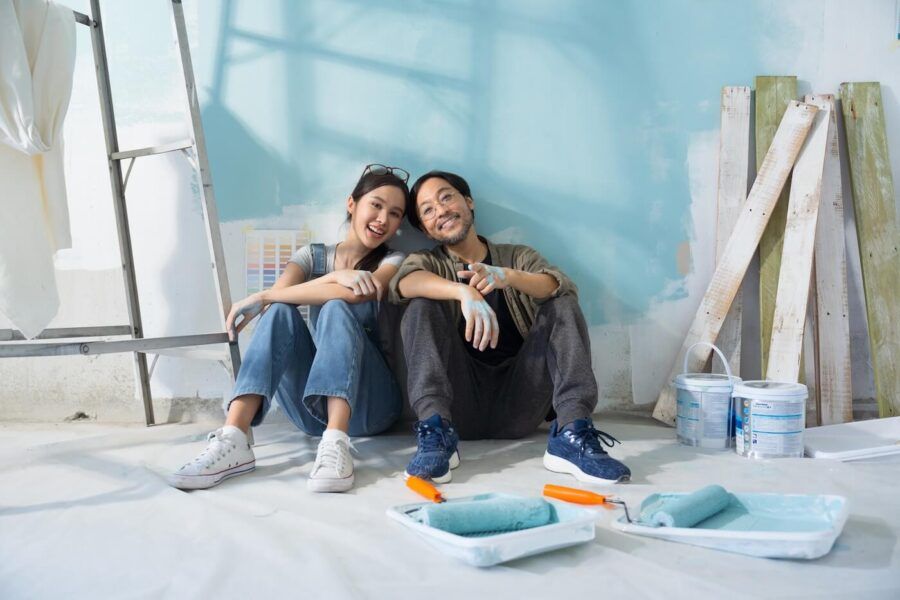 Young couple sitting on the floor taking a break from home renovation. They are sitting next to a ladder, with blue paint cans, rollers, and painting supplies scattered around.