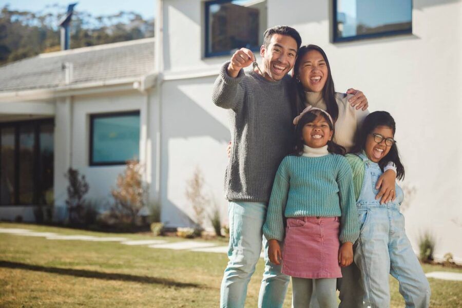 Cheerful family of four standing on a green lawn in front of their new house on a sunny day, proudly holding house keys.