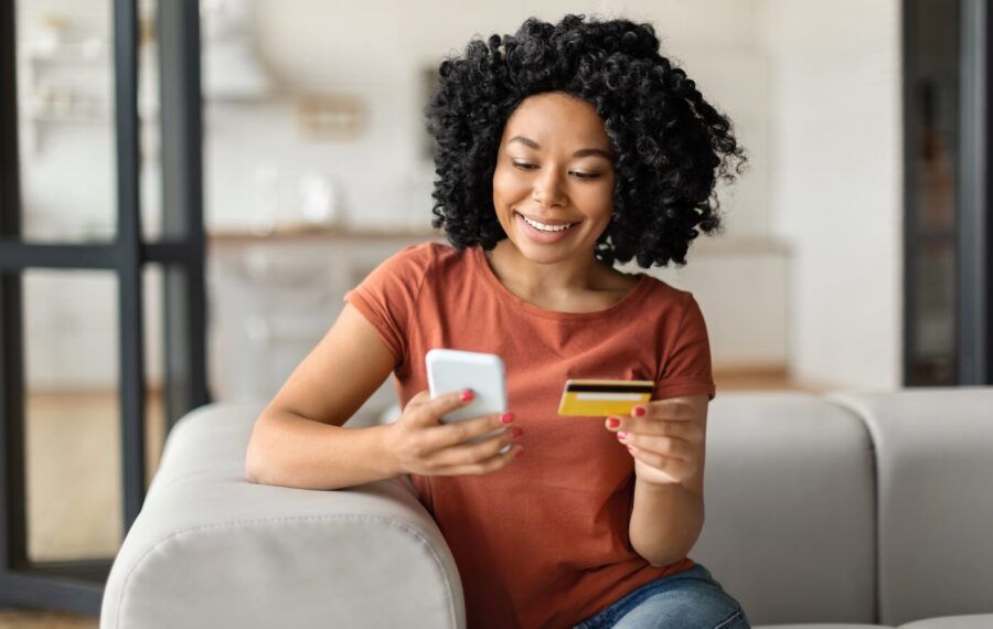 Smiling young woman sitting on a couch abd using her smartphone while holding a credit card