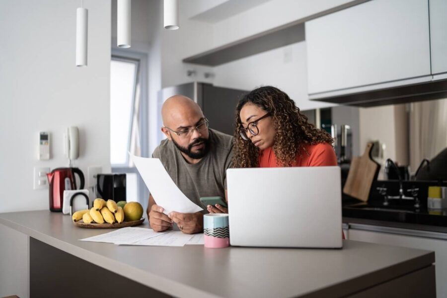 Focused couple reviewing documents together at a kitchen counter, with a laptop, papers, and a bowl of bananas nearby