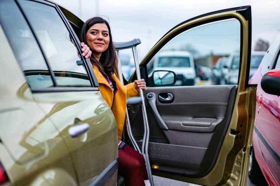 Smiling woman using crutches while stepping out of a car in a parking lot.