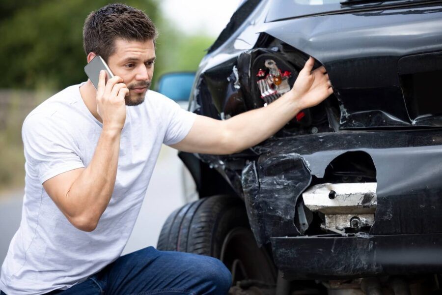 Man squatting next to his damaged car, making a phone call to his auto insurance company