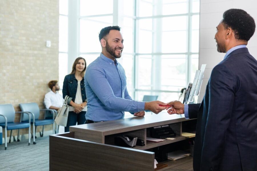 Smiling man giving his debit card to a male clerk at the bank counter, people are standing in the line behind him