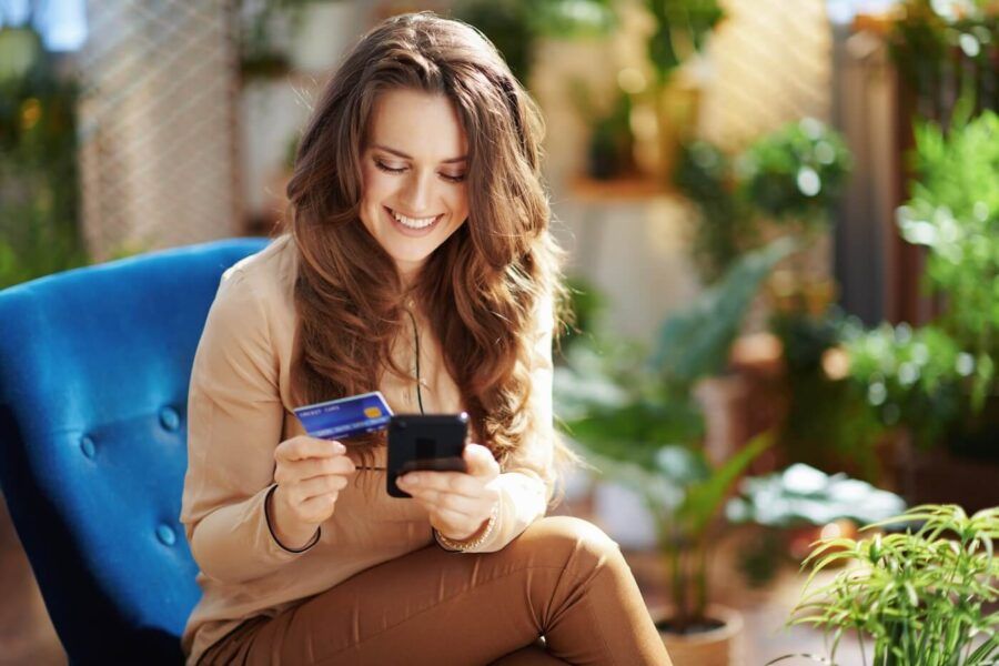 Smiling woman sitting on a blue armchair, paying with a credit card via mobile phone