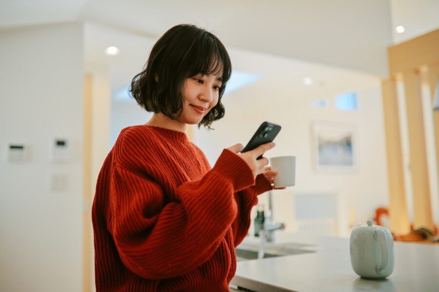 Smiling young woman wearing a red sweater is checking her credit score on her smartphone while holding a mug