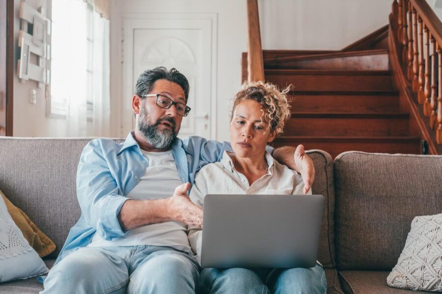 Concerned mature couple sitting on a sofa with a laptop, the man pointing at the laptop screen.