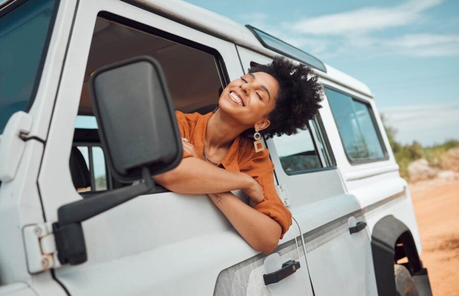 Smiling woman leans out of the open window of a white off‑road vehicle on a sunny day, resting her arms on the door while looking toward the landscape.