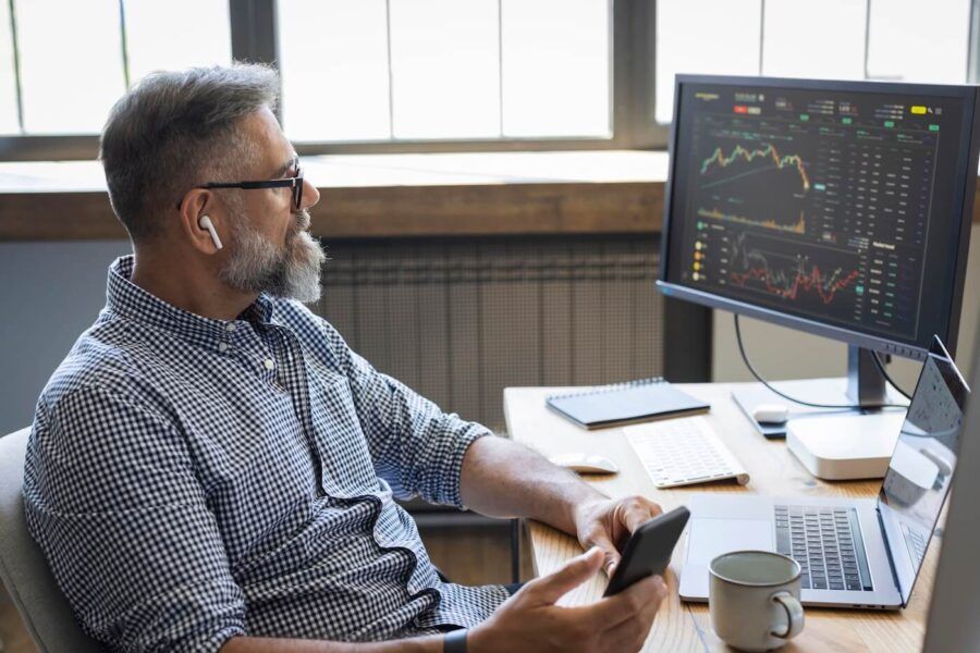 Focused mature man with earbuds sitting at a desk holding a smartphone while looking toward a monitor displaying stock charts and financial data, with a laptop, keyboard, notebook, and coffee mug nearby
