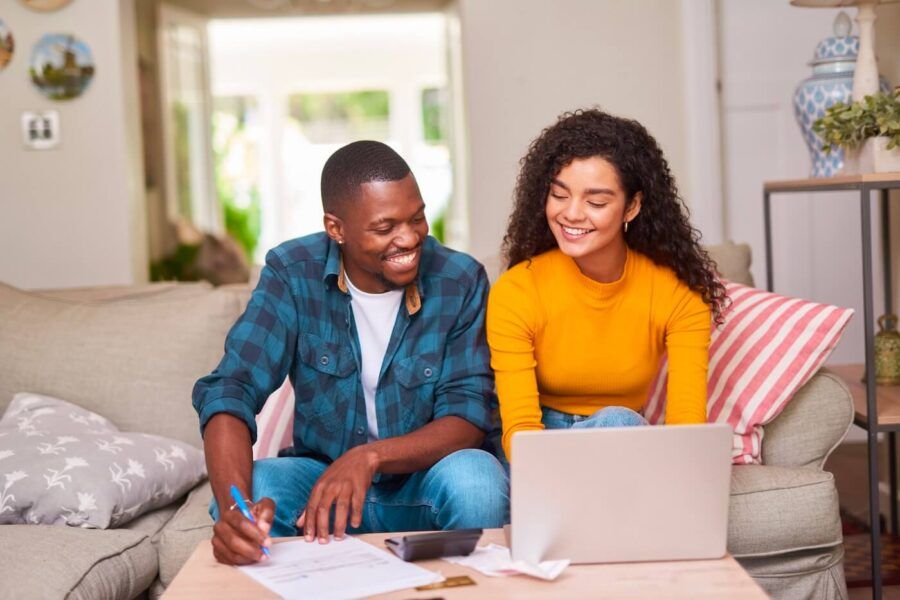 Happy couple sitting together on a couch while reviewing documents at a coffee table, with an open laptop, calculator, and papers spread out in a living room setting.