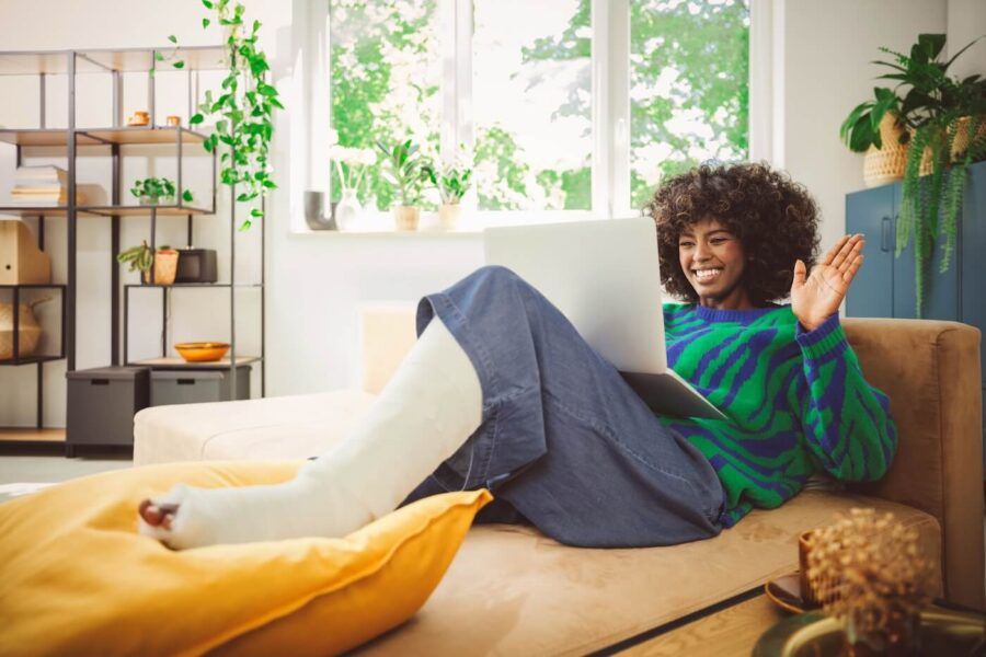 Happy young woman with a casted leg lying on a sofa and having a video call on her laptop