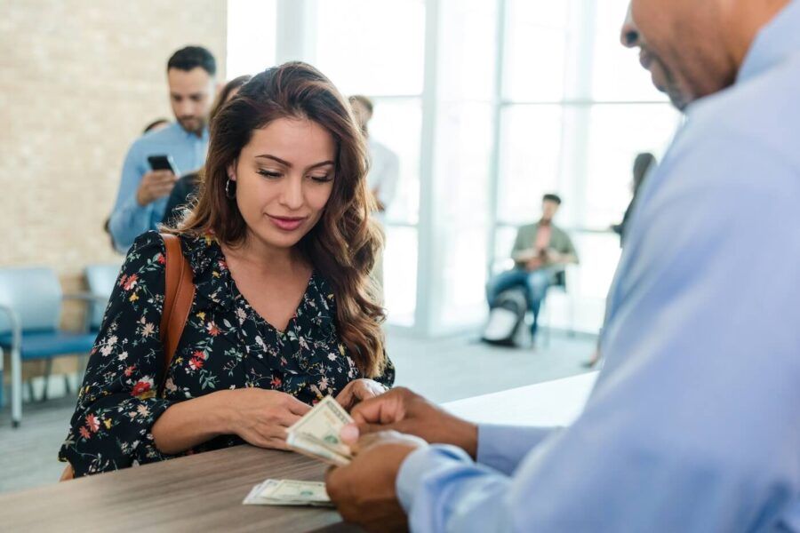 Woman standing at a customer service counter while a bank employee counts and hands over cash. Other customers are visible in the background of a bright financial institution