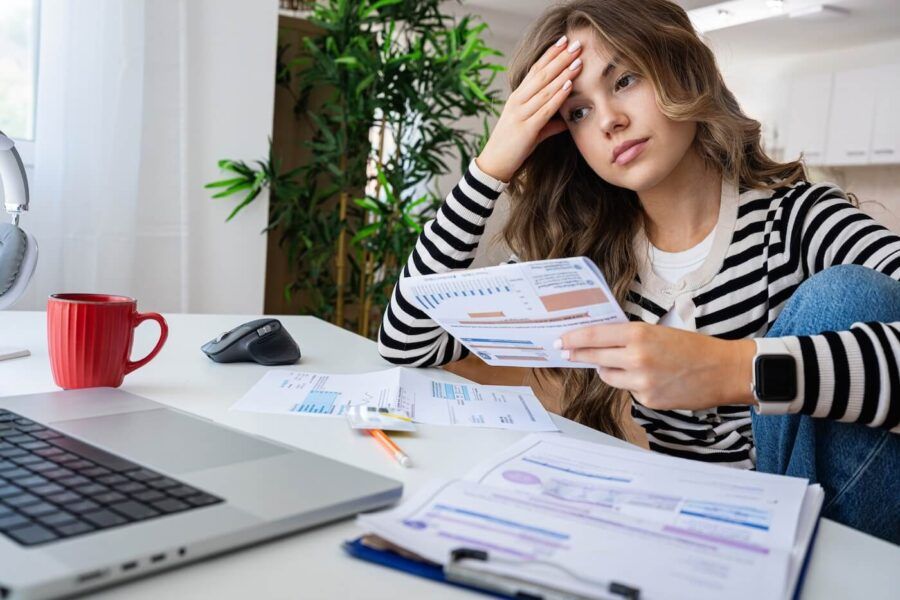 Pensive young woman seated at a desk reviewing financial statements and bills, with a laptop, documents, calculator, and coffee mug nearby.