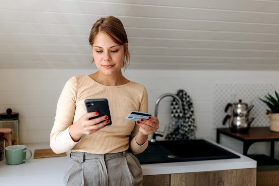 Focused young woman standing in a modern kitchen holding a smartphone in one hand and a credit card in the other. The image illustrates online shopping, mobile banking, or digital payments, with a sink, countertop, mug, and kitchen appliances visible in the background.